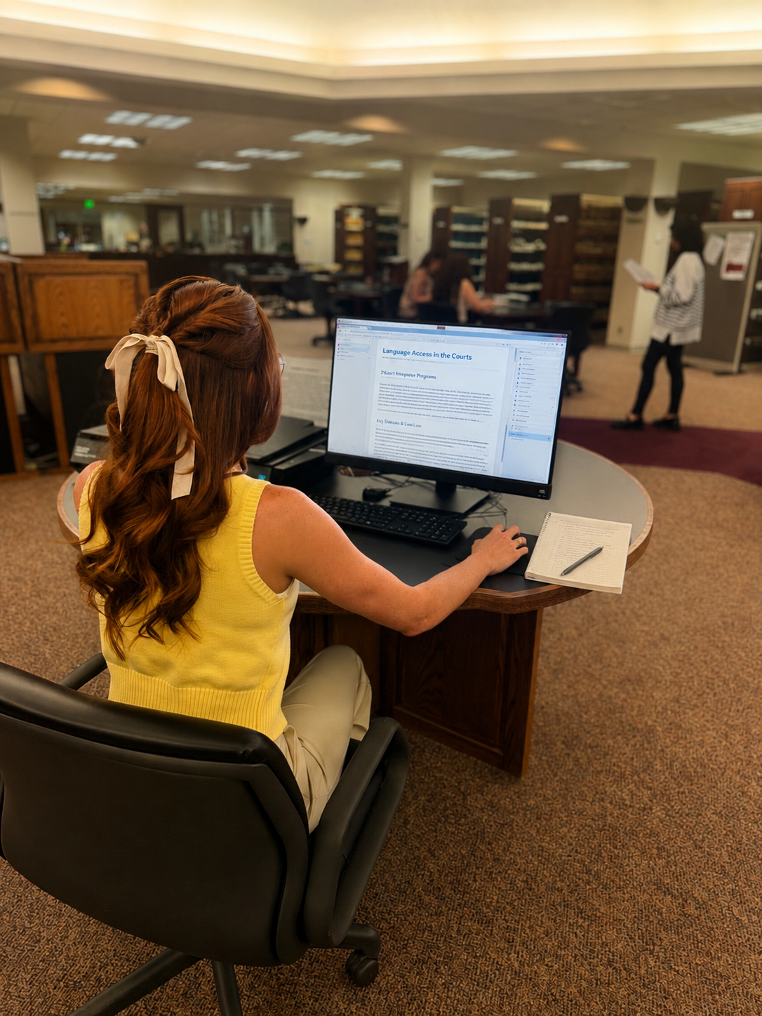 This image features a woman operating a computer showing the usage of the law library with others looking at books behind her desk